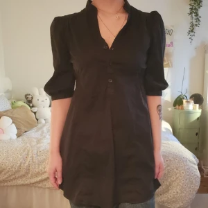 Svart kortklänning med puffärm - Svart kortklänning med puffiga ärmar och v-ringad hals. Knappar framtill och en lätt A-linjeformad siluett.