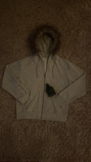 Grå hoodie zip med päls - Grå unisex zip up hoodie med på sydd päls i huvan. Storlek ”s”. Helt ny. Skickar fort