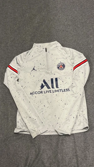 PSG långärmad träningströja Jordan - Snygg PSG träningströja från Jordan i ljusgrått med svarta prickar, lång dragkedja och klubbmärke på bröstet. Röd och svart rand på ärmarna, tillverkad i tekniskt material som andas. Perfekt för dig som gillar fotboll och streetwear.