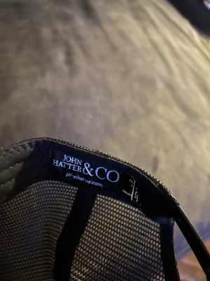 Svart keps John Hatter & Co Sparta - Cool svart keps från John Hatter & Co med mesh på sidorna och bak. Framtill sitter en metallskylt med röd text 'THIS IS SPARTA!' och röd ram. Kepsen har böjd skärm och justerbar passform, perfekt för dig som vill sticka ut.