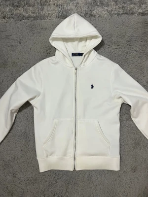  Vit Polo kofta - Vit hoodie från Polo Ralph Lauren med dragkedja och huva. Tröjan har två stora fickor framtill och ett mörkblått broderat logomärke på bröstet. Insidan är mjuk och fleecefodrad, perfekt för kyliga dagar. Klassisk och clean design som funkar till allt.