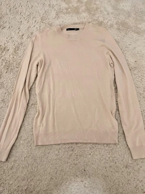 Beige stickad tröja med rund hals - Snygg beige stickad tröja från lager 157 med rund hals och långa ärmar. Tröjan har ribbade muddar vid ärmslut och nederkant, vilket ger en clean och stilren look. Perfekt för dig som gillar enkla och neutrala färger.