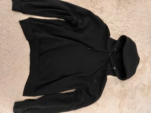 Svart hoodie med huva och snörning - Svart hoodie med klassisk huva och snörning framtill. Tröjan har långa ärmar och ribbade muddar vid ärmslut och nederkant. Perfekt för chill dagar eller när du vill ha en enkel och stilren look.