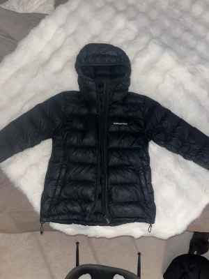 Svart dunjacka Peak Performance - Svart dunjacka från Peak Performance med huva och dragkedja framtill. Jackan har en quiltad design med horisontella paneler och två sidofickor med dragkedja. Perfekt för kalla dagar och har en clean, sportig look. Jackan är i gott skick förutom ett hål på armen, därmed diskuterbart pris. 
