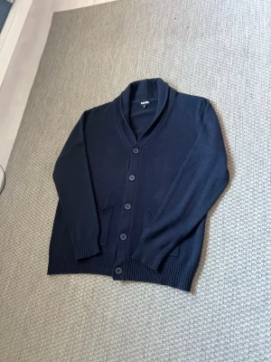 Mörkblå kofta  - Snygg mörkblå kofta från Watsons i storlek XL men passar även L. Koftan har ribbade muddar, två framfickor och stängs med fem knappar. Materialet är stickat och ger en chill och klassisk vibe. Perfekt att slänga på över en t-shirt när det är lite kyligt ute.