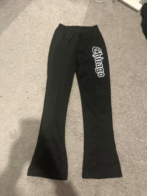 Svarta flared mjukis byxor från chicago. Passar mig som 164 - Svarta bootcut byxor med elastisk midja och stort vitt Chicago-tryck på ena benet. Snyggt utsvängda ben och mjukt material som ger en skön passform. Perfekta för dig som gillar streetstyle och vill sticka ut med coola detaljer.