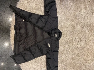 Svart pufferjacka från The North Face - Svart pufferjacka från The North Face i storlek S. Jackan har en klassisk quiltad design, hög krage och dragkedja framtill. Perfekt för kalla dagar med sin fluffiga och isolerande känsla. Logotypen syns tydligt på både bröstet och ryggen. Snygg och enkel att matcha.
