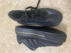 Svarta Adidas sneakers med gulddetaljer - Säljer ett par svarta Adidas sneakers med gulddetaljer på hälen och sidan. Skorna har klassisk snörning, platt sula och rund tå. Materialet är en mix av syntet och skinn, med Adidas-logga i guld på baksidan. Perfekt för dig som gillar stilrena och sportiga skor.