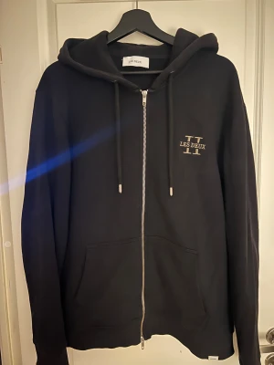 Svart hoodie med dragkedja Les Deux - Svart hoodie från Les Deux med silverfärgad dragkedja, huva och snörning. Tröjan har broderad logga på bröstet och fickor framtill. Perfekt för chill dagar och enkel att matcha med jeans eller joggers.