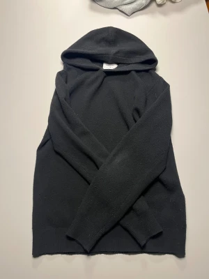 Svart hoodie i merinoull från Vailent - Svart hoodie från Vailent i mjuk merinoull. Tröjan har huva, långa ärmar och ribbade muddar vid ärmslut och nederkant. Perfekt för dig som gillar stilrena och bekväma plagg med lite extra lyxig känsla. Snygg att matcha med jeans eller joggers.