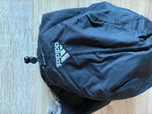 Svart pälsfodrad vinterhatt Adidas - Säljer en svart vinterhatt från Adidas med ClimaProof-tryck. Hatten har pälsfoder i grått och svart, samt justerbar snörning och kardborreband under hakan. Perfekt för kalla dagar när du vill hålla dig varm och ändå se sportig ut.