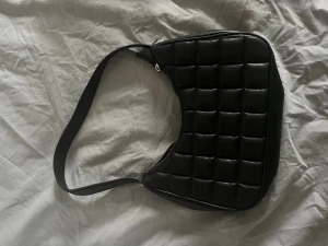 Svart quiltad axelväska i skinn - Snygg svart axelväska med quiltad design och rektangulär form. Väskan har en bred axelrem och stängs med dragkedja upptill. Materialet är skinn och insidan är fodrad i svart tyg. Perfekt storlek för det viktigaste och ger en cool vibe till din outfit. Har några defekter vid slutet av remmarna.