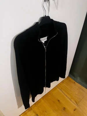 Svart stickad kofta från Maison Margiela - Svart stickad kofta från Maison Margiela med ribbad struktur och hel dragkedja framtill. Tröjan har lång ärm, hög krage och klassiska vita stygn i nacken. Perfekt för dig som gillar minimalistisk och stilren design. Använd 1 gång( med tags på) finns spår av parfym doft. Mvh ItsForfits 