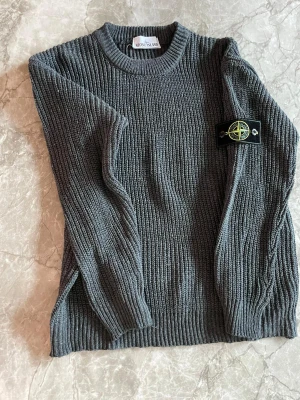Grå stickad tröja från Stone Island - Säljer en grå stickad tröja från Stone Island med klassisk rund hals och ribbade muddar. Tröjan har lång ärm och den ikoniska Stone Island-patchen på vänster arm. Perfekt för dig som gillar streetwear och snygga detaljer.
