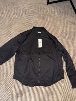 Svart overshirt från cp company - Svart overshirt från cp company med lång ärm och klassisk krage. Framtill finns en bröstficka och en hel dragkedja. På ena ärmen sitter en rund detalj som ger en cool touch. Perfekt för dig som gillar stilrena och funktionella plagg.