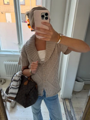 Beige stickad kofta med kort ärm - Säljer en beige stickad kofta med korta ärmar och bred krage. Koftan har snyggt kabelstickat mönster, knappar framtill och ribbade detaljer vid kragen och ärmsluten. Perfekt att slänga över en t-shirt för en chill look.