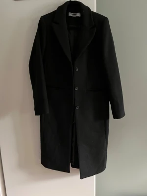 Svart lång kappa med knappar - Säljer en svart, stilren och lång kappa med klassisk krage och tre knappar framtill. Modellen är rak och har diskreta fickor på sidorna. Perfekt för dig som gillar minimalistisk och clean look. Materialet känns som ullblandning och kappan har lång ärm.