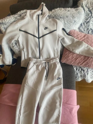 Vit Nike tech set - Säljer en fräsch vit hoodie från Nike med hel dragkedja och huva. Tröjan har svarta detaljer och logga på bröstet, samt långärmad design. Materialet är mjukt och känns som bomull, perfekt för chill eller träning. Snyggt sportigt snitt och ribbade muddar.