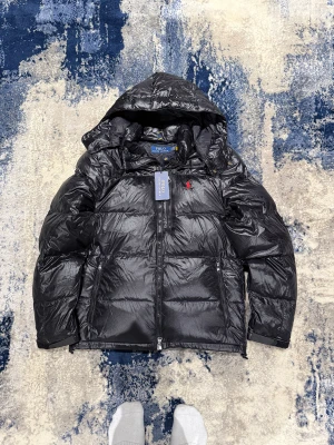 Polo Ralph Lauren vinterjacka - Säljer en svart dunjacka från Polo Ralph Lauren med huva och blank finish. Storlek L men passar även M. Perfekt för kalla dagar och riktigt snygg streetstil.