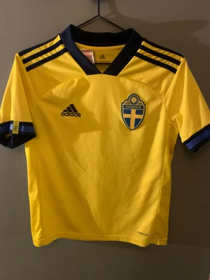 Svenska landslagets gulblå fotbollströja - Gul fotbollströja från Adidas med Sveriges landslagsemblem på bröstet och svarta Adidas-ränder på axlarna. Blå detaljer vid krage och ärmslut, samt texten 'Sverige' på ryggen. Tillverkad i lätt, ventilerande syntetmaterial med Aeroready-teknologi.