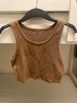 Brun croppad linnetopp i bomull - Säljer en brun croppad linnetopp med grov ribbad struktur. Toppen är armlös och har rundad halsringning. Materialet är mjukt och känns skönt mot huden, perfekt för varma dagar. Enkel och stilren design som är lätt att matcha med jeans eller shorts.