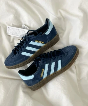 Adidas Spezial blå sneakers mocka - Säljer ett par Adidas Spezial sneakers i mörkblå mocka storlek 41, med ljusblå och ränder på sidorna. Skorna har bruna gummisulor, snörning och rund tå. Guldtexten 'Spezial' syns på sidan. Perfekt för dig som gillar retrostil och klassiska streetwear-vibbar. Dessa skor är även helt oanvända!