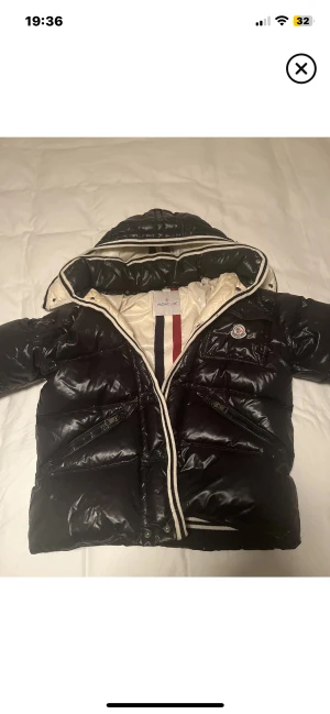 Svart Moncler pufferjacka med huva - Svart pufferjacka från Moncler med glansig finish, huva och vita detaljer längs dragkedjan. Jackan har en bröstficka med Moncler-logga, två sidofickor med dragkedja och quiltad design. Insidan är ljus med röd och marinblå rand. Perfekt för kalla dagar.