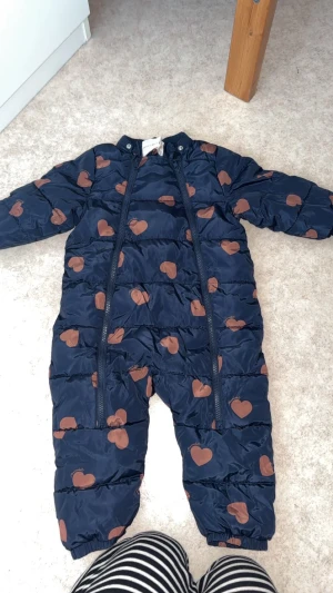 Mörkblå overall med hjärtan - Säljer en mörkblå overall  i storlek 86, täckt med bruna hjärtan. Jackan har två dragkedjor framtill och är helfodrad för extra värme. Perfekt för kalla dagar och har en gullig design som sticker ut.