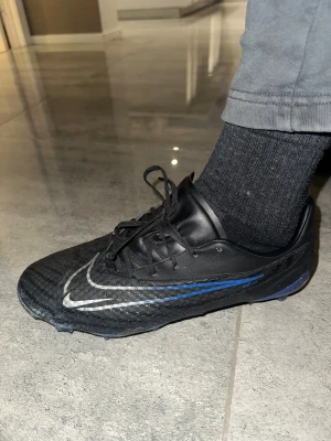 Nike Phantom svart/blå fotbollsskor - Nike Phantom fotbollsskor i svart med blå detaljer och silverfärgad swoosh. Skorna har snörning, platt sula med dobbar och en rund tå. Ovandelen är tillverkad i syntetmaterial för bra grepp och känsla på planen. Perfekt för dig som vill ha stil och funktion på träningen.