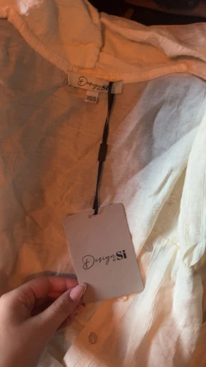 Ljusbeige skjorta från Design by Si - Säljer en ljusbeige skjorta från Design by Si med v-ringad omlottfront och knappar. Skjortan har långa ärmar med lite puff. Den är helt oanvänd lappen sitter kvar på.