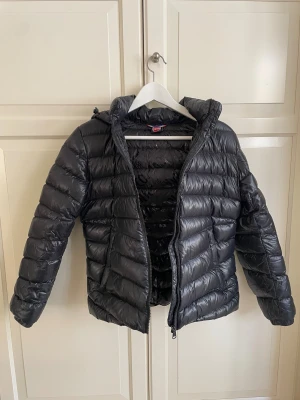 Svart dunjacka från Colmar - Svart dunjacka med huva från Colmar med glansig finish och tryckt logomönster på insidan. Jackan är i fint skick, inga skador i skalet. Har en lagning på fickans insida, se bild nr 6. Perfekt för kalla dagar och har en modern, puffad look som är riktigt snygg.