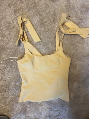 Ljusgul linnetopp med knytband - Supersöt ljusgul linnetopp med breda axelband som knyts i rosetter på axlarna. Toppen har en fyrkantig halsringning och är figurnära i modellen. Perfekt att styla med jeans eller kjol för en fräsch look.