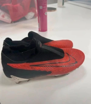 Nike fotbollsskor svart/röd - Säljer ett par grymma Nike fotbollsskor i svart och rött med stor Swoosh-logga på sidan. Skorna har en strömlinjeformad design, platt sula med dobbar och är tillverkade i syntetmaterial med mesh för bra ventilation. Perfekt för dig som vill ha stil på planen.
