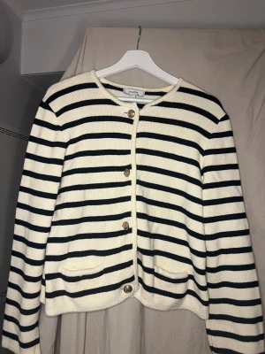Randig kofta från Rihoas i S - Snygg stickad kofta från Rihoas i storlek S. Den är vit med svarta horisontella ränder, har guldfärgade knappar framtill och två små fickor. Perfekt för dig som gillar klassisk och chill stil.