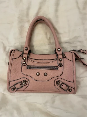 Rosa väska med nitar - Bra skick men några defekter, dragkedjan på framsidan har tappat sitt snöre (men den funkar fortfarande) snöret till dragkedjan ovanpå väskan har stretchats ut lite, och svarta kanten på handtaget har lossnat lite. Allt syns på bilderna. Det är inga stora defekter som är så märkbara så väskan är fin ändå! Och det ingår även ett längre band till väskan som man kan ta av och på.