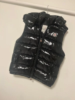 Svart dunväst från Moncler med huva - Snygg svart dunväst från Moncler med glansig finish och avtagbar huva. Västen har två dragkedjeförsedda fickor, quiltad design och Moncler-logga på bröstet. Perfekt för lager-på-lager och streetstyle. Stängs med dragkedja framtill.