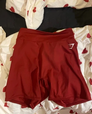 Röda gymshark shorts - Säljer ett par röda Gymshark shorts med hög midja och snygg scruch baktill. De är tillverkade i ett stretchigt syntetmaterial som sitter tight och är perfekta för träning. Loggan syns tydligt på sidan och färgen är djup röd. Storleken är L men passar S-L