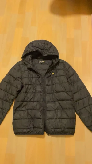 Svart pufferjacka från Lyle & Scott - Svart pufferjacka från Lyle & Scott med huva och gul logga på bröstet. Jackan har quiltad design, dragkedja framtill och är tillverkad i syntetmaterial. Perfekt för kyliga dagar och har en clean, modern look som funkar till streetwear.