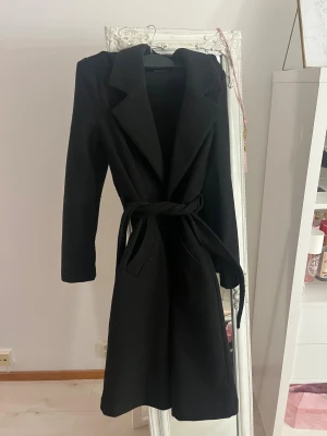 Svart kappa från Vero Moda XS - Svart lång kappa från Vero Moda i klassisk modell med bred krage och knytskärp i midjan. Snygg och stilren med långa ärmar och två fickor framtill. Perfekt för dig som gillar en clean och tidlös look.