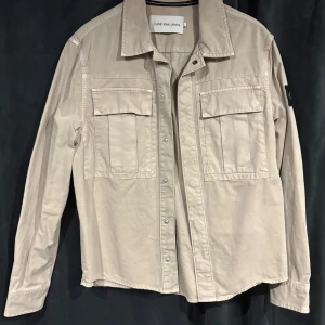 Beige overshirt från Calvin Klein Jeans - Stilren beige overshirt från Calvin Klein Jeans med klassisk krage, två stora bröstfickor med lock och tryckknappar framtill. Långärmad modell med svart logopatch på ena ärmen. Perfekt lager-på-lager-plagg i mjuk bomull. Aldrig använd då jag råkade köpa den i förliten storlek. Storlek S