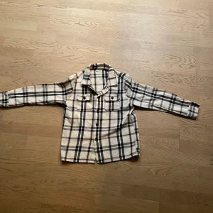 Rutig skjorta i beige och svart - Snygg rutig skjorta i beige och svart med klassisk krage och två bröstfickor med knappar. Skjortan har långa ärmar och stängs med knappar framtill. Perfekt för en avslappnad och trendig look.