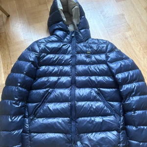 Blå dunjacka med huva från Blauer - Säljer en mörkblå dunjacka från Blauer med huva och dragkedja. Jackan har två snedställda fickor med dragkedja, quiltad design och Blauer-logga på bröstet och ärmen. Perfekt för kalla dagar och riktigt snygg streetstil.