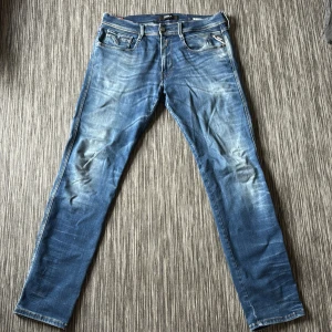 Replay Anbass Hyperflex jeans blå 33 - Säljer ett par Replay Anbass Hyperflex jeans i klassisk blå tvätt med snygga slitningar. Modellen har normal passform och raka ben, med fem fickor och knappgylf. Stretchigt material för extra komfort och rörelsefrihet.