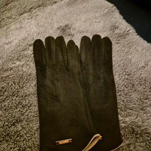 Svarta mockahandskar med metallmärke - Snygga svarta handskar i mjuk mocka med stilren design och ett litet metallmärke med texten 'Fashion' vid handleden. Handskarna har en klassisk form med fem fingrar och en diskret ljus kantdetalj vid öppningen. Perfekta för dig som gillar enkel och elegant stil.