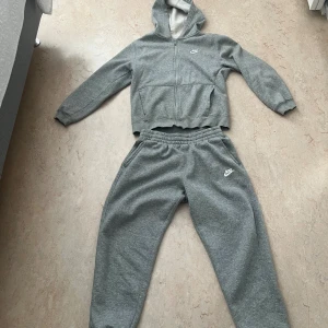 Grå mjukisbyxor från Nike XL(barn)=S - Gråa mjukisbyxor från Nike i storlek XL med broderad vit logga på benet. Byxorna har resår och snörning i midjan samt mjuk fleece på insidan för extra komfort. Perfekta för chill eller träning.