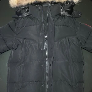 Svart dunjacka från Canada Goose - Säljer en svart dunjacka från Canada Goose med päls på huvan och klassisk logga på ärmen. Jackan har dragkedja och knappar framtill, flera fickor och glansigt svart innerfoder. Perfekt för kalla vinterdagar och riktigt snygg streetstil.