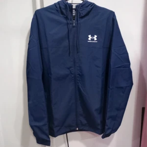Blå Under Armour - Snygg marinblå vindjacka från Under Armour med huva och dragkedja framtill. Jackan har meshfoder, två fickor med dragkedja och logga på bröstet. Perfekt för träning eller när du vill ha en sportig look.