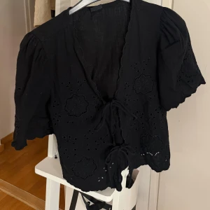 Svart blus med broderi från Gina Tricot - Supersöt svart blus från Gina Tricot med broderade blommor och knytning framtill. Blusen har korta puffärmar och v-ringning, samt fina hålbroderier och vågiga kanter.