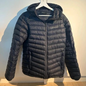 Jacka  Peak Performance - Snygg Mörkblå dunjacka från Peak Performance med huva och dragkedja framtill. Jackan har quiltad design och är lätt vadderad, perfekt för kyliga dagar. Klassisk passform och diskret logga på bröstet. Enkelt stilren och passar till det mesta.