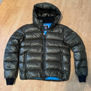 Rossignol grön dunjacka  - Säljer en mörkgrön dunjacka från Rossignol med huva och svarta muddar. Jackan har blått foder, två fickor med dragkedja. Perfekt nu inför de kalla dagarna. Vid fler frågor är det bara att höra av sig!🙌Ny pris: 5000kr